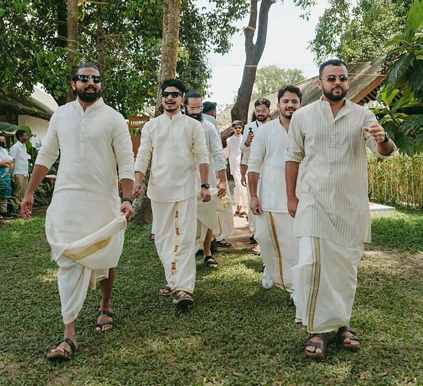 Men walking across a lawn in ivory kurtas and gold-bordered mundus